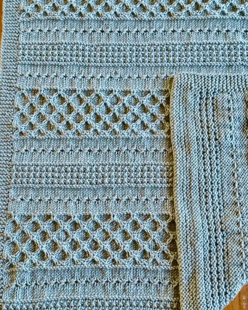 May include: A close-up of a light blue knitted blanket with various textured patterns. The blanket features rows of horizontal ribbing, diamond-shaped openwork, and a textured border. The corner of a smaller, matching square is visible.
