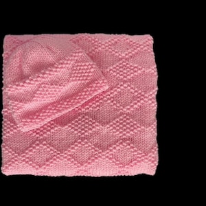 Op de afbeelding: Een gebreide baby set in zachtroze, bestaande uit een muts en een deken. De deken heeft een textuur van ruitpatroon. De set is gemaakt van een zacht, warm materiaal, ideaal voor een pasgeborene.