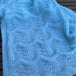 May include: A light blue knitted blanket with a detailed pattern. The blanket has a textured design with a scalloped edge. The soft, cosy blanket is suitable for a baby or as a decorative throw.