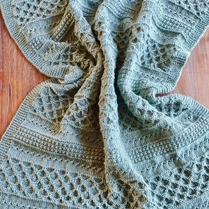 May include: A sage green knitted blanket with intricate openwork and textured patterns. The blanket is draped over a wooden surface, showcasing its detailed design and soft texture. The knitted design features geometric and floral patterns.
