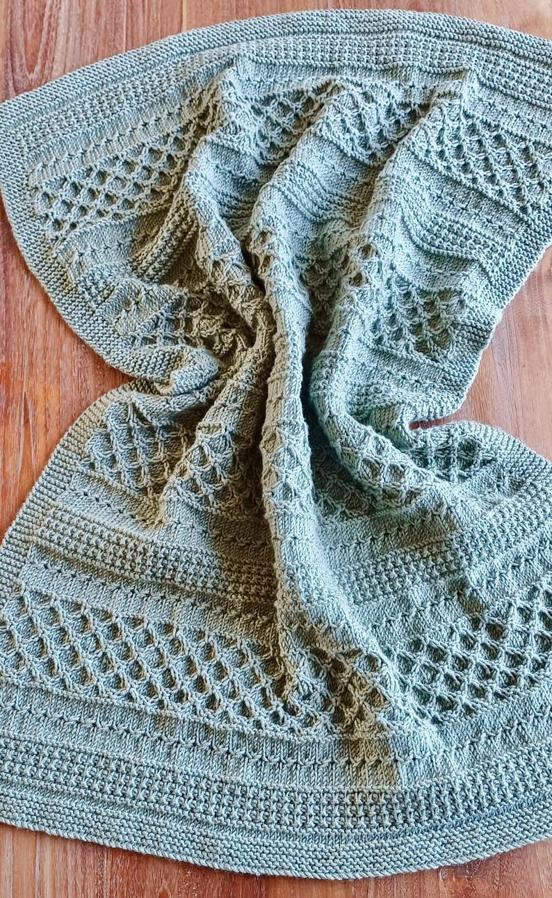 May include: A sage green knitted blanket with intricate openwork and textured patterns. The blanket is draped over a wooden surface, showcasing its detailed design and soft texture. The knitted design features geometric and floral patterns.