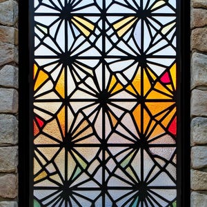 Puede incluir: Una ventana de vitrales con un patrón geométrico de líneas negras y vidrio de colores. El vidrio es en tonos de amarillo, naranja, rojo, azul y verde.