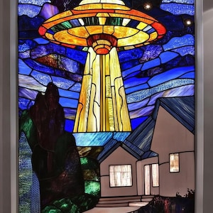 Puede incluir: Una ventana de vitral que representa un platillo volante sobrevolando una casa con un camino que lleva a la puerta principal. El cielo es azul con nubes blancas y estrellas. El platillo es amarillo, naranja y rojo. La casa es blanca con un techo marrón. El camino es gris.