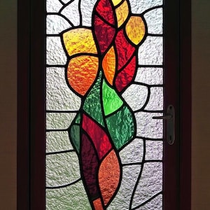 Puede incluir: Una ventana de vitrales con un diseño abstracto colorido que presenta piezas de vidrio rojo, naranja, amarillo y verde. El diseño se asemeja a una flor o planta estilizada.