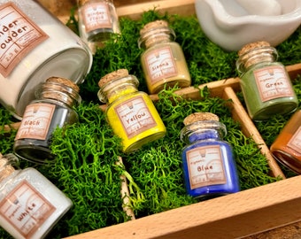 May include: A set of small glass bottles with cork stoppers, filled with different colored pigments. The bottles are labeled with the pigment color, such as "Black", "White", "Yellow", "Blue", "Green", and "Sienna". The bottles are arranged in a wooden box with moss lining the bottom.