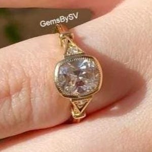 May include: A gold ring with a square, clear gemstone. The ring has a detailed, vintage-inspired design with smaller accent stones. The text "GemsBySV" is visible on the image.