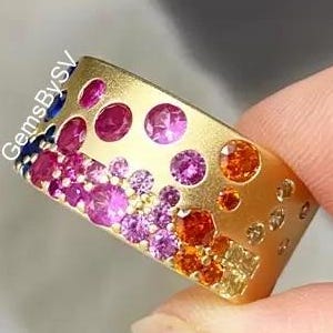 Peut inclure: Une bague dorée ornée d'un dégradé de pierres précieuses colorées. La bague présente un mélange de pierres roses, rouges, oranges et jaunes, ainsi que quelques pierres bleues. La bague a une surface texturée.