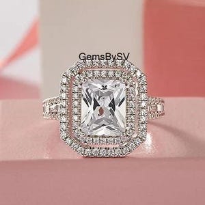 Könnte beinhalten: Ein silberner Ring mit einem großen, rechteckigen, klaren Edelstein in der Mitte, umgeben von einem doppelten Halo aus kleineren, klaren Steinen. Der Ring liegt auf einer rosa Oberfläche, mit einem verschwommenen Hintergrund.