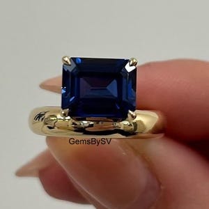 May include: A gold ring featuring a large, square-cut, deep blue gemstone. The ring's band is a smooth, rounded design. The text "GemsBySV" is visible on the ring.