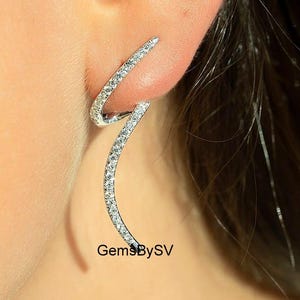 Può includere: Orecchini in argento dal design curvo e aperto, incastonati con piccole pietre scintillanti. Il marchio "GemsBySV" è visibile. Gli orecchini sono indossati su un orecchio.