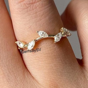 Puede incluir: Un anillo de oro con una banda curva adornada con cinco piedras claras de talla marquesa dispuestas en forma de hoja. La banda de metal tiene un acabado pulido y el texto GemsBySV es visible en pequeñas letras negras en el centro.