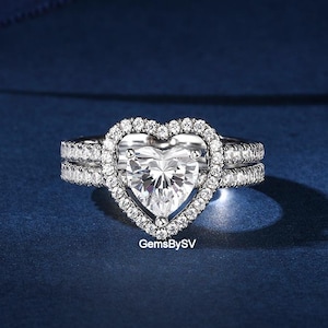 May include: A heart-shaped diamond ring with a halo of smaller diamonds. The ring sits on a band with additional diamond accents. The background is a deep blue, and the text "GemsBySV" is visible.