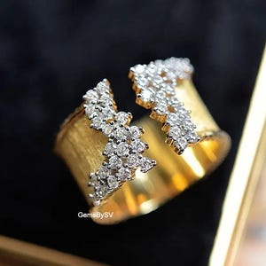 Puede incluir: Un anillo dorado con un diseño dividido, adornado con grupos de pequeños diamantes brillantes. El anillo tiene una banda texturizada y está sobre un fondo oscuro. El texto "GemsBySV" es visible.