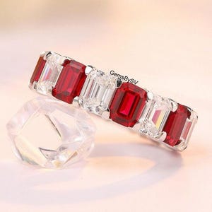 Puede incluir: Un anillo plateado con gemas rectangulares rojas y transparentes alternas. El anillo está sobre una superficie rosa claro, con una gema clara y facetada en primer plano. El texto "GemsBySV" es visible.