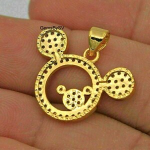 May include: A gold-toned pendant shaped like a circular head with two round ears, featuring a smaller circular head design in the center. The surface is covered in small, circular indentations and includes a bail at the top. Text reads GemsBySV.