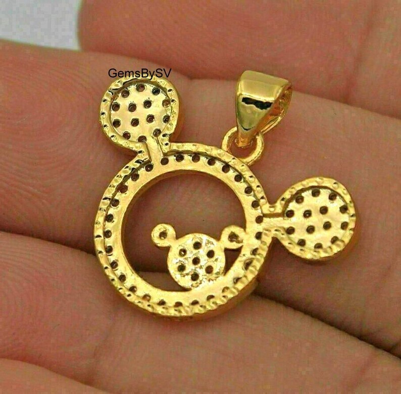 May include: A gold-toned pendant shaped like a circular head with two round ears, featuring a smaller circular head design in the center. The surface is covered in small, circular indentations and includes a bail at the top. Text reads GemsBySV.