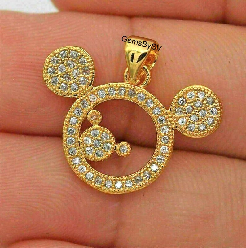May include: A gold-toned pendant shaped like a circular mouse head silhouette, featuring a large central ring and two smaller circular ears, all encrusted with clear pav&eacute;-set stones. The text GemsBySV is printed in black near the top bail.