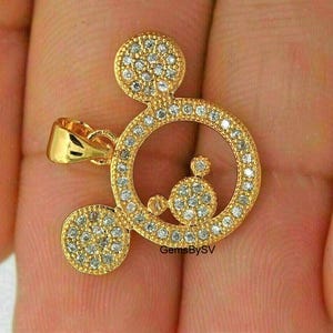 May include: A gold-toned pendant shaped like a circular silhouette with two smaller circular ears, all encrusted with small clear stones. The pendant features a polished gold bail on the left side and includes the text GemsBySV printed on the surface.