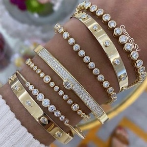 Op de afbeelding: Verschillende gouden armbanden met diamantaccenten worden getoond op een pols. De armbanden variëren in ontwerp, waaronder een rij diamanten, een band met verspreide diamanten en een band met diamanten. De tekst "GemsBySV" is zichtbaar.