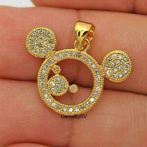 May include: A gold-toned pendant shaped like a circular head with two smaller circular ears, featuring a pav&eacute; setting of clear, sparkling stones throughout the entire surface. The pendant includes a small bail at the top for attachment.