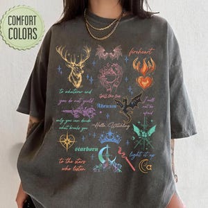 May include: A charcoal grey short-sleeve t-shirt featuring a multi-coloured graphic print of a deer, dragon, crown, and various mystical symbols. The design includes text such as "Fireheart," "Abraxos," "Starborn," and "Hello Witchling" alongside small stars.