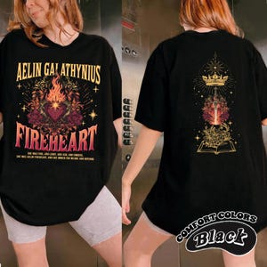 Może przedstawiać: Czarny t-shirt z grafiką "AELIN GALATHYNIUS FIREHEART". Przedni wz&oacute;r przedstawia serce, płomienie i elementy kwiatowe. Tył ma koronę, miecz i wz&oacute;r książki. "COMFORT COLORS Black" jest wydrukowane w prawym dolnym rogu.