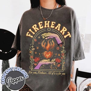 May include: A charcoal grey t-shirt featuring a graphic print of two hands cupping a flaming heart within an arched frame. The text reads "FIREHEART" at the top and "For you, Fireheart. All of it is for you" at the bottom, surrounded by floral accents.