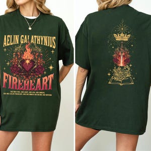 Może przedstawiać: Ciemnozielona koszulka z napisem "AELIN GALATHYNIUS FIREHEART" w kolorze złotym i czerwonym. Prz&oacute;d zdobi serce, płomienie i elementy kwiatowe. Tył zdobi korona, miecz i wz&oacute;r otwartej książki.
