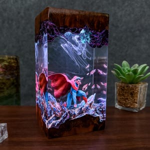 Puede incluir: Escultura rectangular de resina con Superman en una pose dinámica. La escultura está encapsulada en resina transparente con una parte superior oscura con aspecto de madera. La escena incluye una capa roja, un traje azul y una base rocosa detallada, con un efecto brillante.