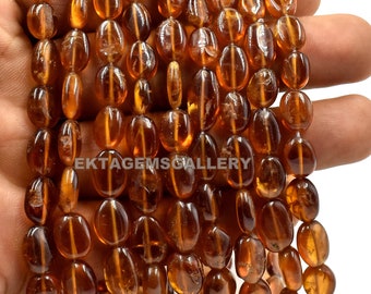 May include: A strand of oval-shaped, translucent, orange-brown gemstone beads. The beads are smooth and polished, with a slight sheen. The beads are strung on a clear thread.