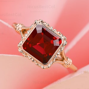 May include: A gold ring with a square-shaped red gemstone set in a delicate, ornate setting. The ring is photographed on a pink background.