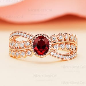May include: A rose gold ring with a large, round, deep red gemstone at the center. The band features a leaf-like design with small, clear gemstones and a delicate, curved shape. The ring is set against a soft pink background.