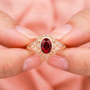 May include: A gold ring featuring a central oval-cut deep red gemstone. The setting is crafted with an intricate filigree design surrounding the stone, accented by small metal beads. The band displays a detailed floral pattern on both sides.
