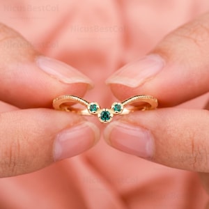 Può includere: Un anello in oro a forma di chevron con tre piccole pietre preziose rotonde di colore verde incastonate al centro. La fascia presenta un dettaglio bordato e testurizzato lungo la parte superiore e inferiore, conferendo un aspetto raffinato.
