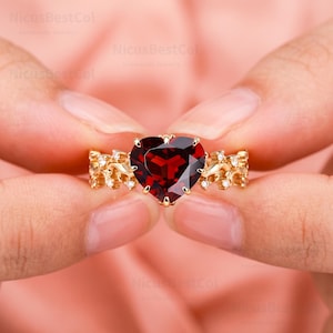 May include: A gold ring featuring a large, heart-shaped, deep red garnet gemstone at the center. The band is crafted with an intricate, vine-like metal design and is accented with small, clear, round-cut stones set along the sides of the band.