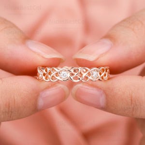 May include: A rose gold band ring featuring an intricate, repeating openwork knot pattern. The metal band is set with small, clear, round-cut stones embedded at regular intervals within the continuous, interconnected, swirling metal design.