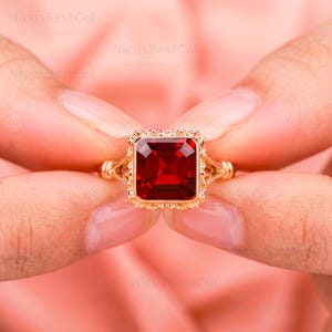 May include: A gold ring featuring a large, rectangular, faceted red gemstone set in a decorative, scalloped metal frame. The band has a split design where it meets the setting. The text NicusBestCol is visible in the background.