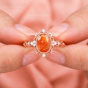 May include: A rose gold ring featuring an oval-cut orange sunstone center gem surrounded by a decorative filigree frame and small round white accent stones on the band. The metal setting has a polished finish and intricate scrollwork details.
