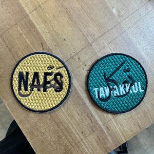 Könnte beinhalten: Zwei runde, gestickte Aufnäher. Einer ist gelb mit schwarzem Text "NAFS" und einem schwarzen Rand. Der andere ist türkis mit weißem Text "TAVAKKUL" und einem schwarzen Rand. Beide liegen auf einer Holzoberfläche.