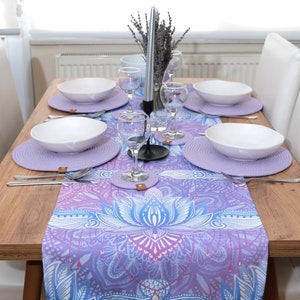May include: A wooden dining table set with a purple and blue patterned table runner. The table is set with white plates, cutlery, and wine glasses. There are four white chairs around the table.
