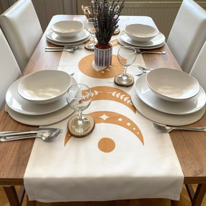 Op de afbeelding: Een witte tafelrunner met een bruin en wit maan- en sterrendesign. De runner ligt op een houten tafel met witte borden, kommen en wijnglazen. In het midden van de tafel staat een vaas met lavendelbloemen.