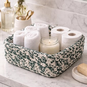 May include: A green and white woven basket contains rolled white towels, toilet rolls, and a lit candle in a glass jar. The basket is on a marble countertop, suitable for a bathroom.