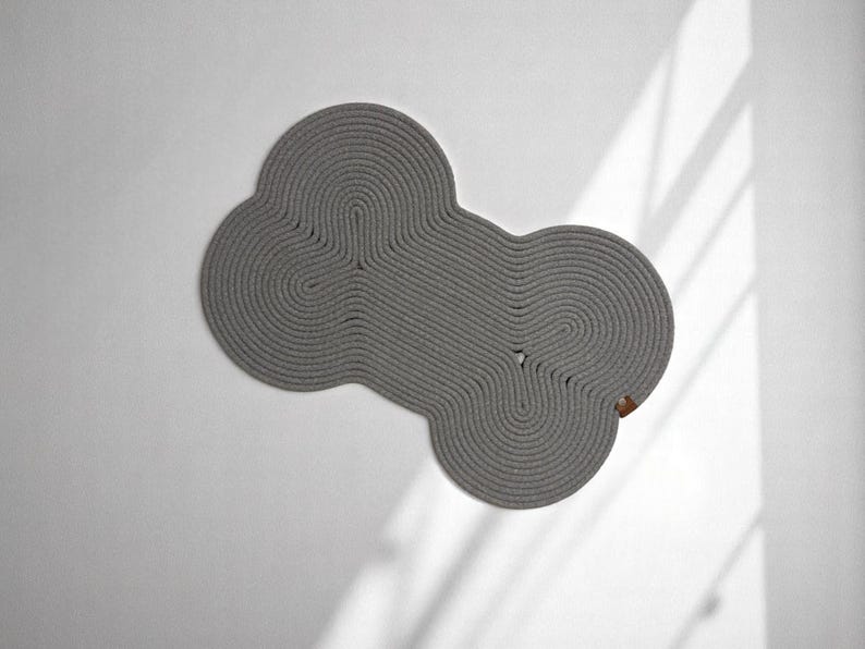 May include: A gray, round, woven rug with a unique, organic shape. The rug is made of thick, rope-like material and has a subtle texture.