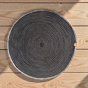 May include: A round, dark gray placemat with a spiral design. The placemat has a white border and a small orange tag. The placemat is made of a woven material and is sitting on a wooden surface.