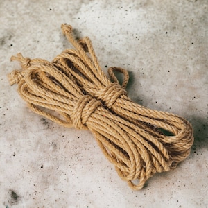 May include: A bundle of natural jute rope, in a tan hue, rests on a speckled grey surface. The rope is tightly coiled and knotted, with several loose ends. This durable rope is suitable for various crafting and decorative projects.