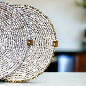 May include: Two round woven placemats with a gold and silver rope design. The placemats have a brown leather tag with the word "Made" on it.