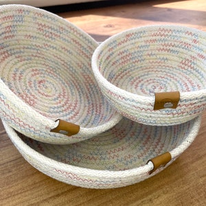May include: Three nested woven rope baskets with a white background and colourful stitching. The baskets have brown leather handles with metal rivets.