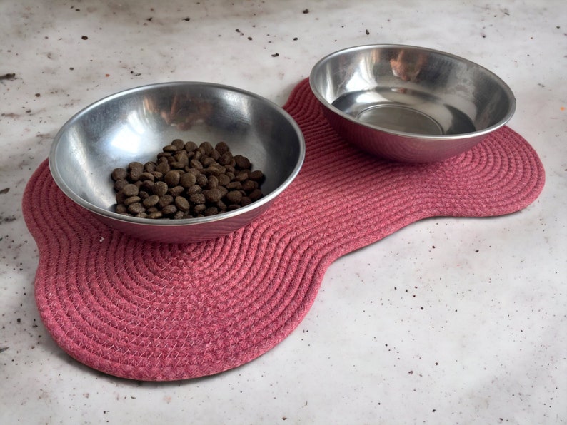 May include: Two stainless steel pet bowls on a pink, rope-like mat. One bowl contains brown dog food.