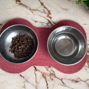 May include: Two stainless steel pet bowls on a pink woven mat. One bowl is filled with brown dog food.
