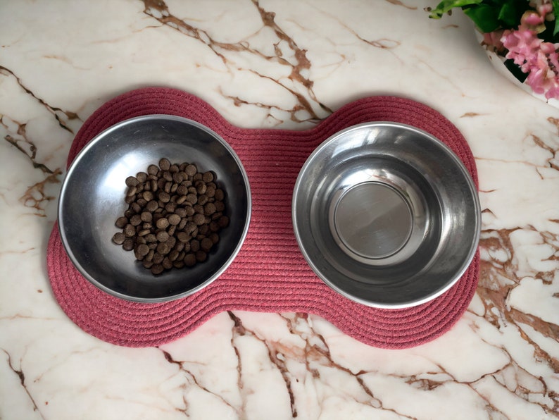 May include: Two stainless steel pet bowls on a pink woven mat. One bowl is filled with brown dog food.